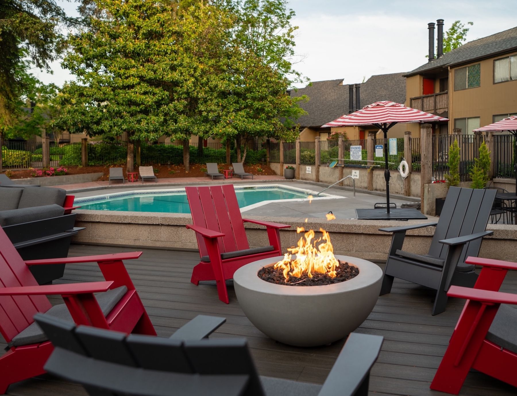 Northgreen Apartments Red and black chairs around a fire pit on a patio, with a pool and striped umbrellas in the background.