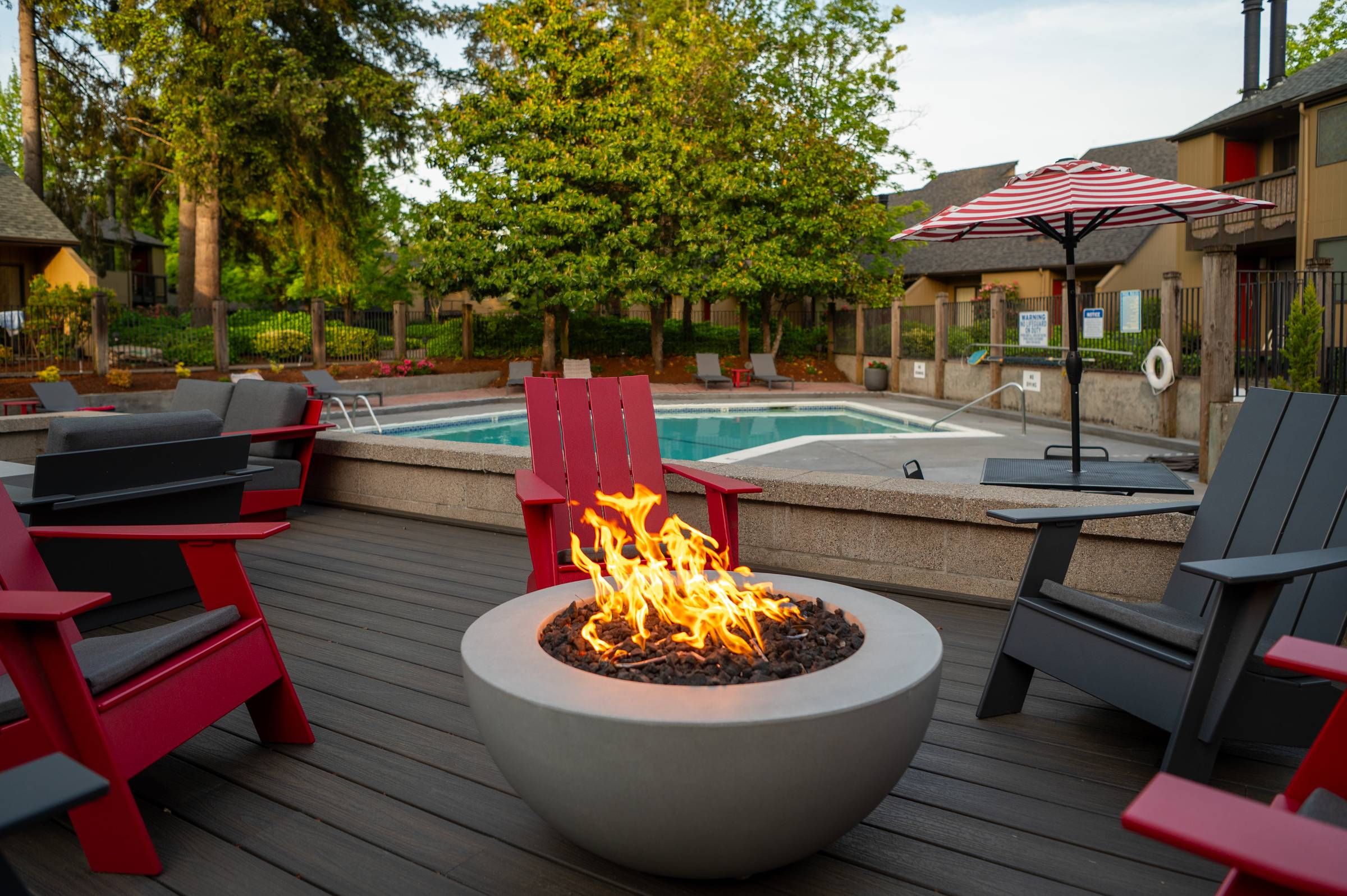 Northgreen Apartments Red and gray chairs surround a round fire pit near a pool with trees and an umbrella in the background.