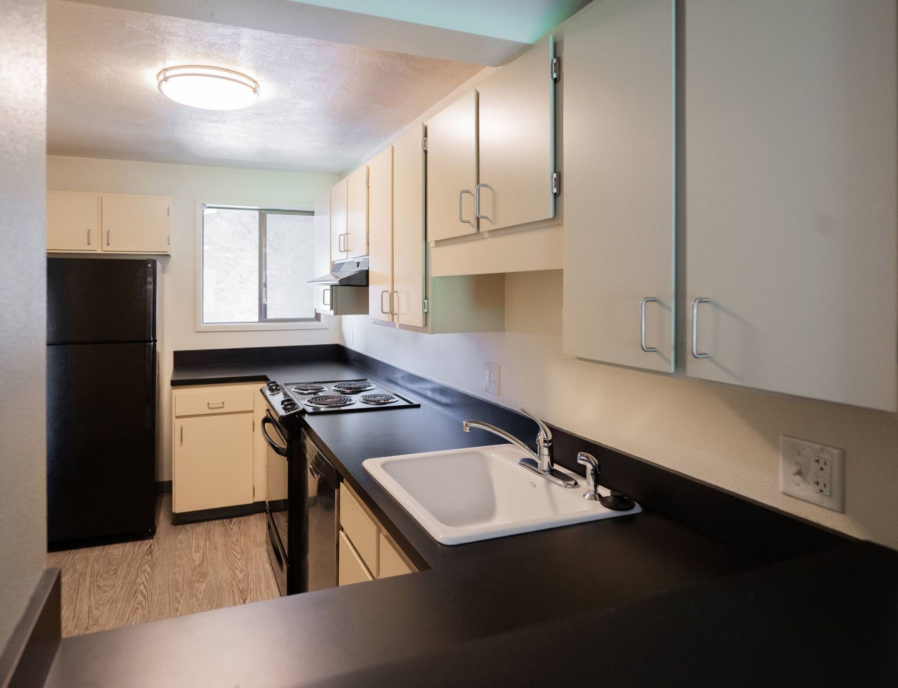Northgreen Apartments Small kitchen with cream cabinets, black appliances, a white sink, and a window letting in natural light.