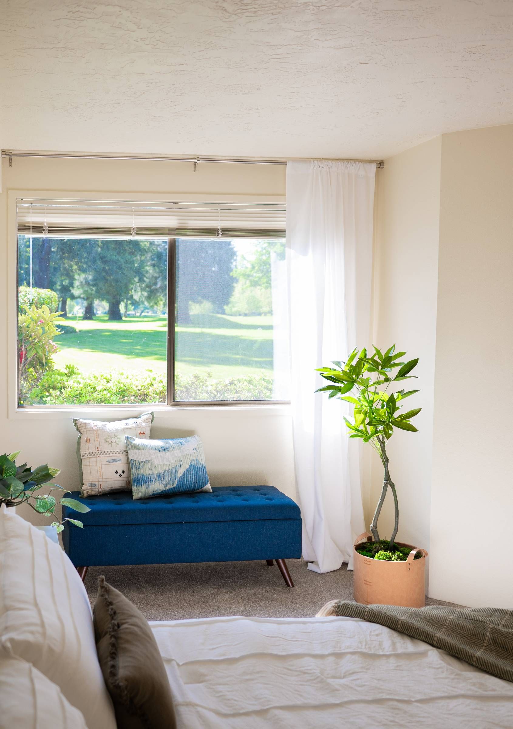 Northgreen Apartments Bright bedroom corner with a blue bench, potted plant, and large window overlooking greenery.