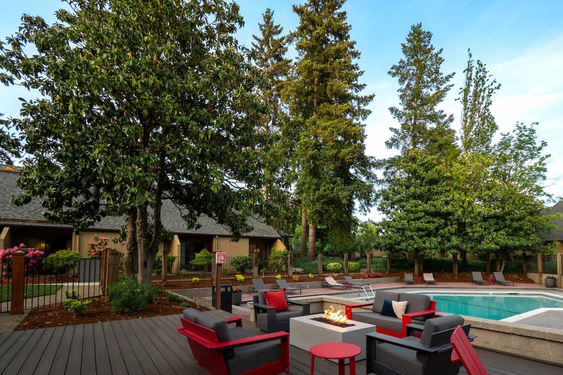 Northgreen Apartments Outdoor patio with red and black chairs around a fire pit, near a swimming pool and surrounded by tall trees.